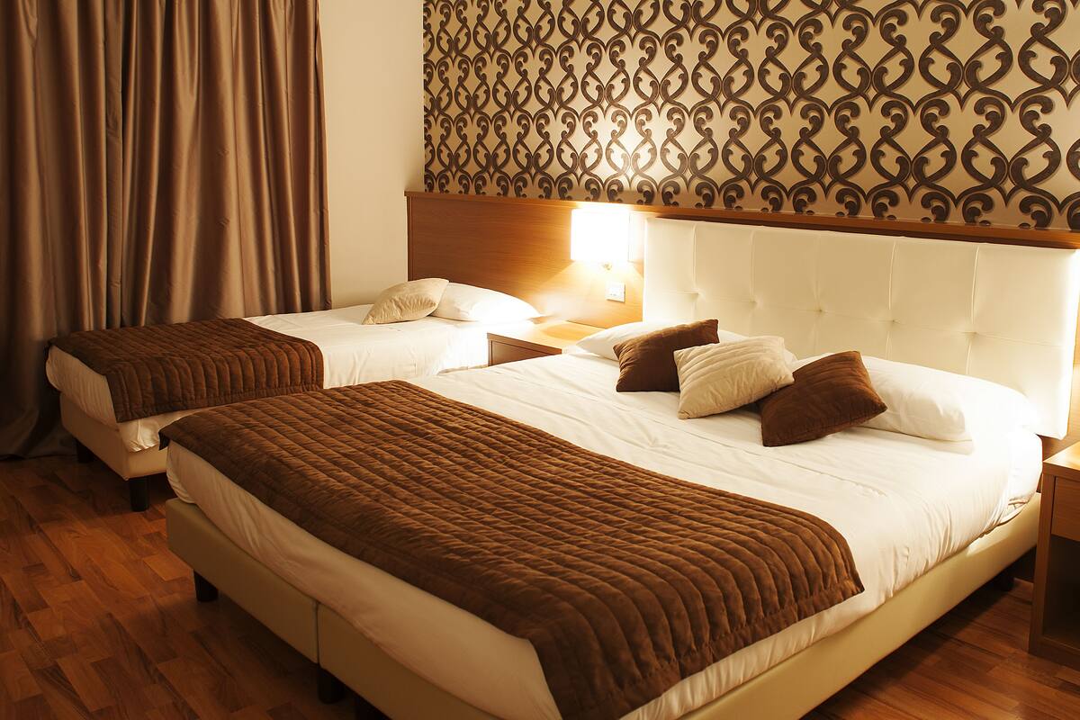 Habitación de hotel con dos camas, suelo de madera, pared con diseño y una mesita de noche con lámpara.