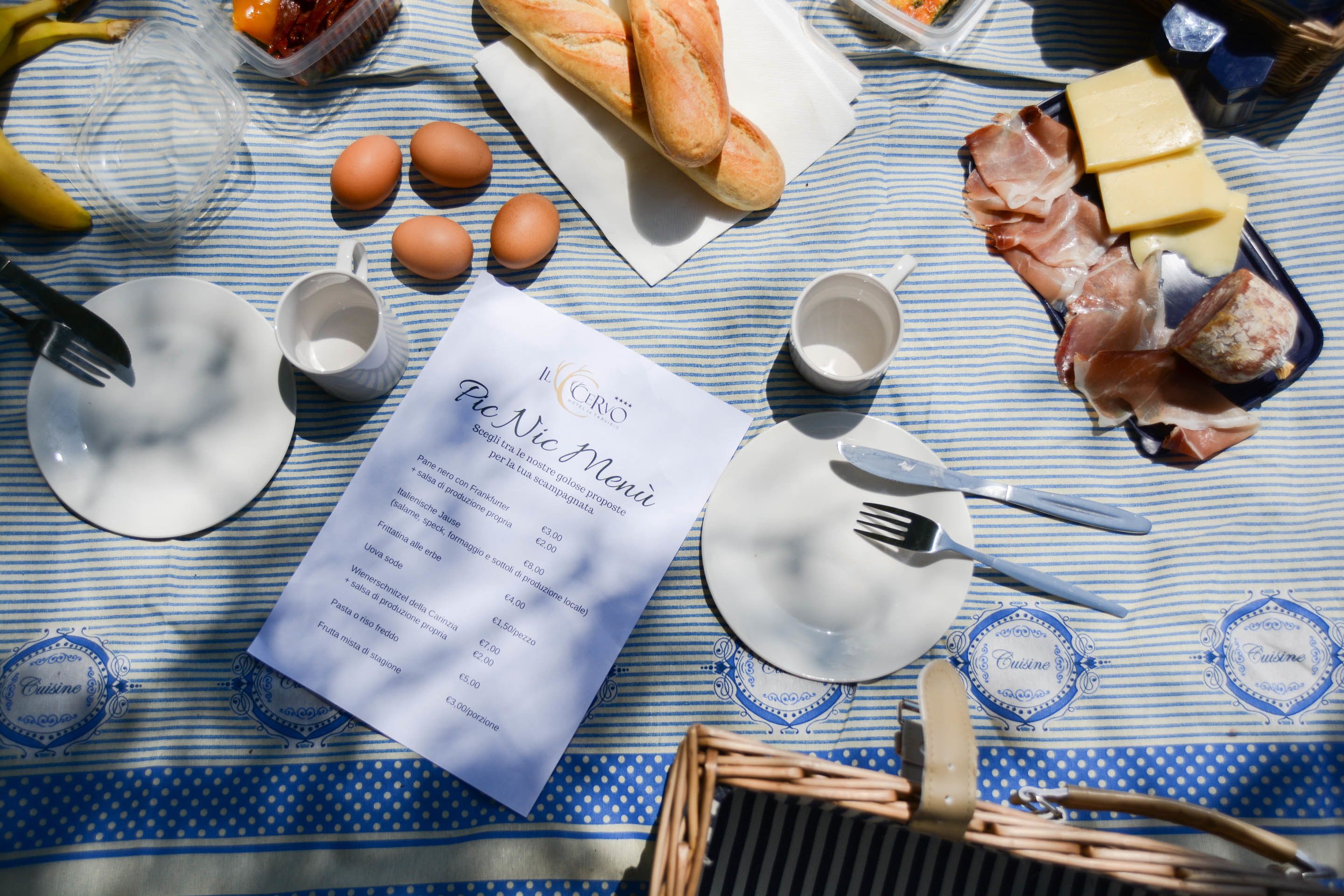 A picnic setup with a menu, bread, eggs, cheese, and meats on a striped tablecloth.