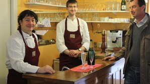 Drei Personen stehen an der Bar, im Hintergrund sind Regale mit Gläsern und Flaschen zu sehen.