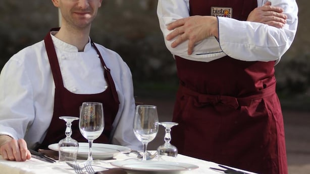 Zwei Kellner an einem Tisch mit weißer Tischdecke und einem Bistro-Sonnenschirm.