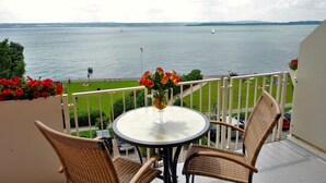 Ein Balkon mit Korbstühlen, einem runden Tisch mit Blumenvase und Blick auf ruhiges Wasser mit Booten.