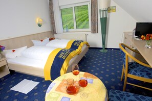 A hotel room with a bed, a desk, a chair, a table with a pitcher and glasses, and a window with a view of green hills. - Seehotel Off (Meersburg)