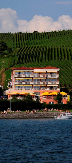 A multi-story hotel with balconies, located by a body of water with people and boats. - Seehotel Off (Meersburg)