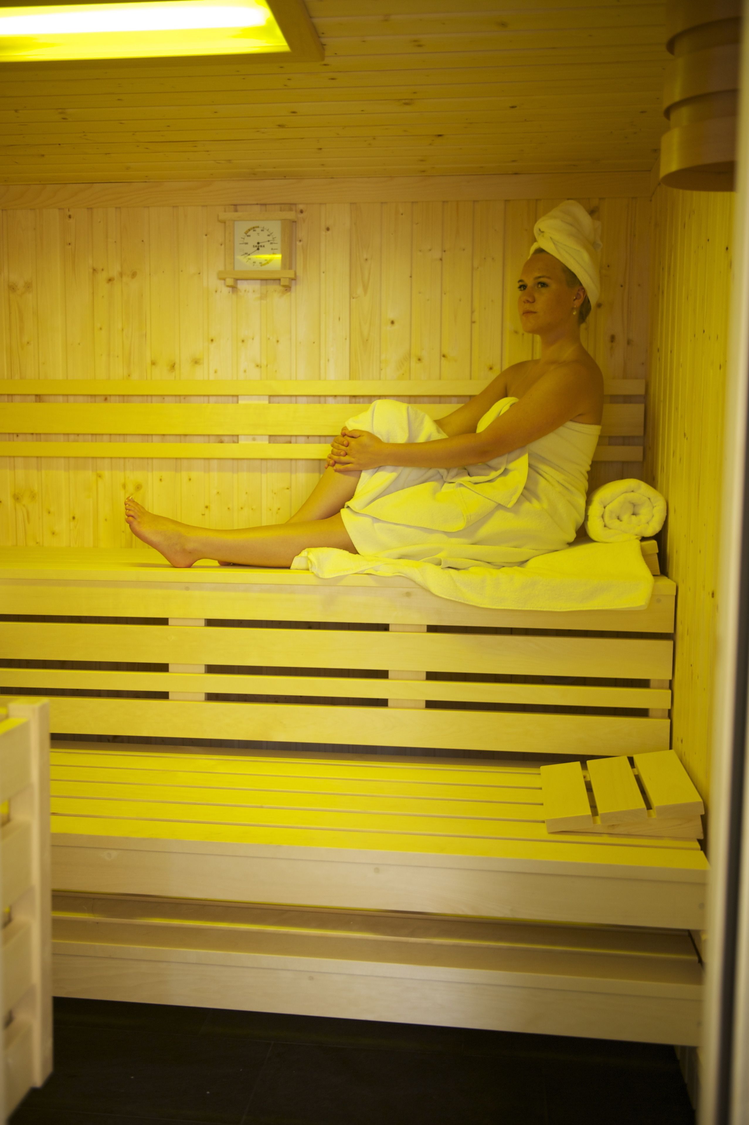 A person sitting in a wooden sauna with a bench and a small clock on the wall.