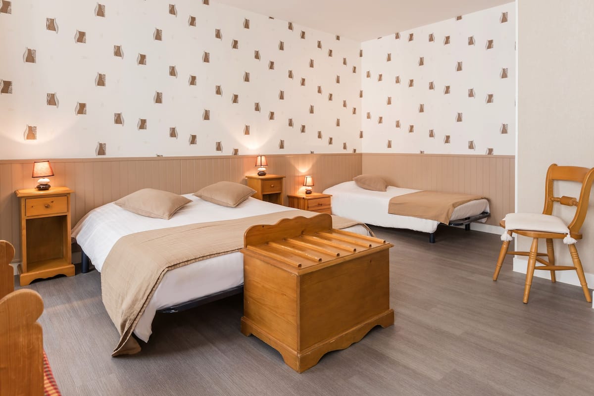 A hotel room with two beds, wooden headboards, bedside tables with lamps, a wooden bench, and a wooden chair.