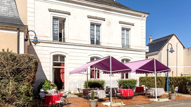 A white building with a balcony, outdoor seating with red and white tablecloths, and purple umbrellas.