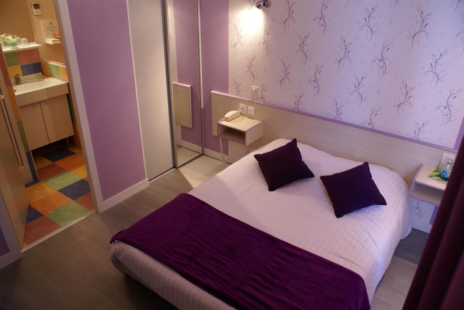 A hotel room with a bed, a nightstand, a mirror, and a bathroom visible through an open door.