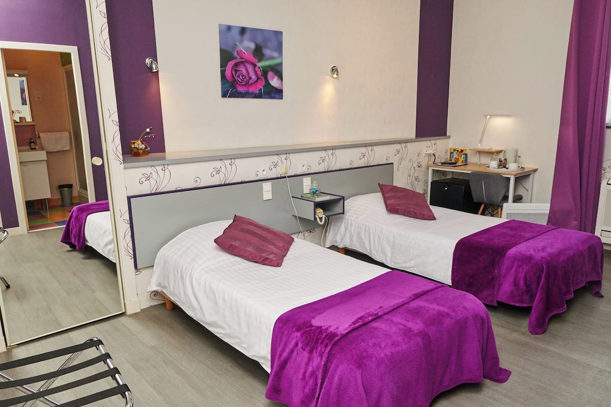 A hotel room with two beds, purple bedding, a desk with a lamp, and a picture of a rose on the wall.