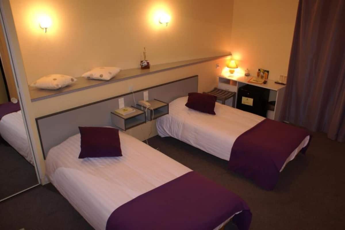 A hotel room with two beds, a bedside table with a lamp, a mirror, and a desk with a chair.