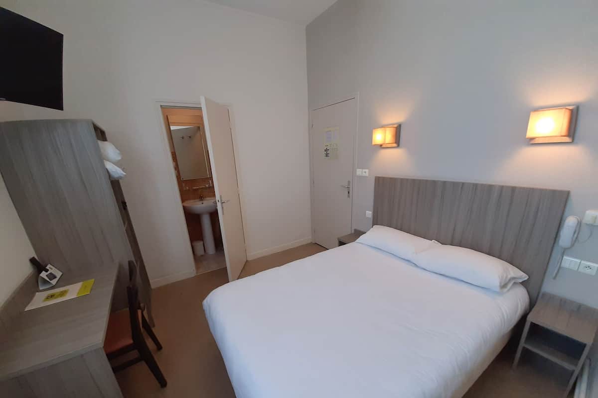 A hotel room with a bed, a desk, a chair, a bathroom visible through an open door, and two wall-mounted lights.