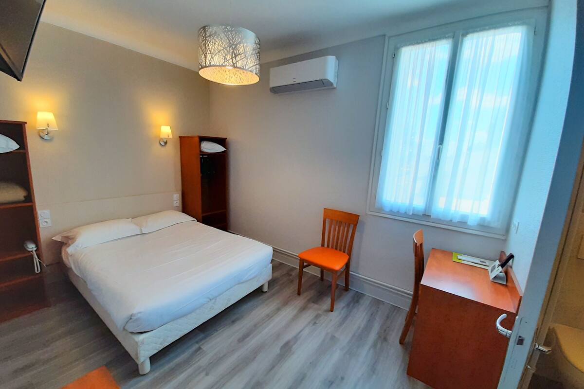 A hotel room with a bed, a desk, a chair, a window with curtains, an air conditioning unit, and a hanging light fixture.