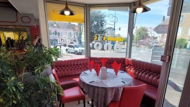 Um restaurante com área de estar acolchoada em vermelho, uma mesa redonda com toalha branca e grandes janelas com vista para a rua.