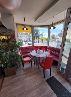 A restaurant with a red cushioned seating area, a round table with a white tablecloth, and large windows offering a view of the street. - Logis hôtel de la Barque (Beaumont-sur-Sarthe)