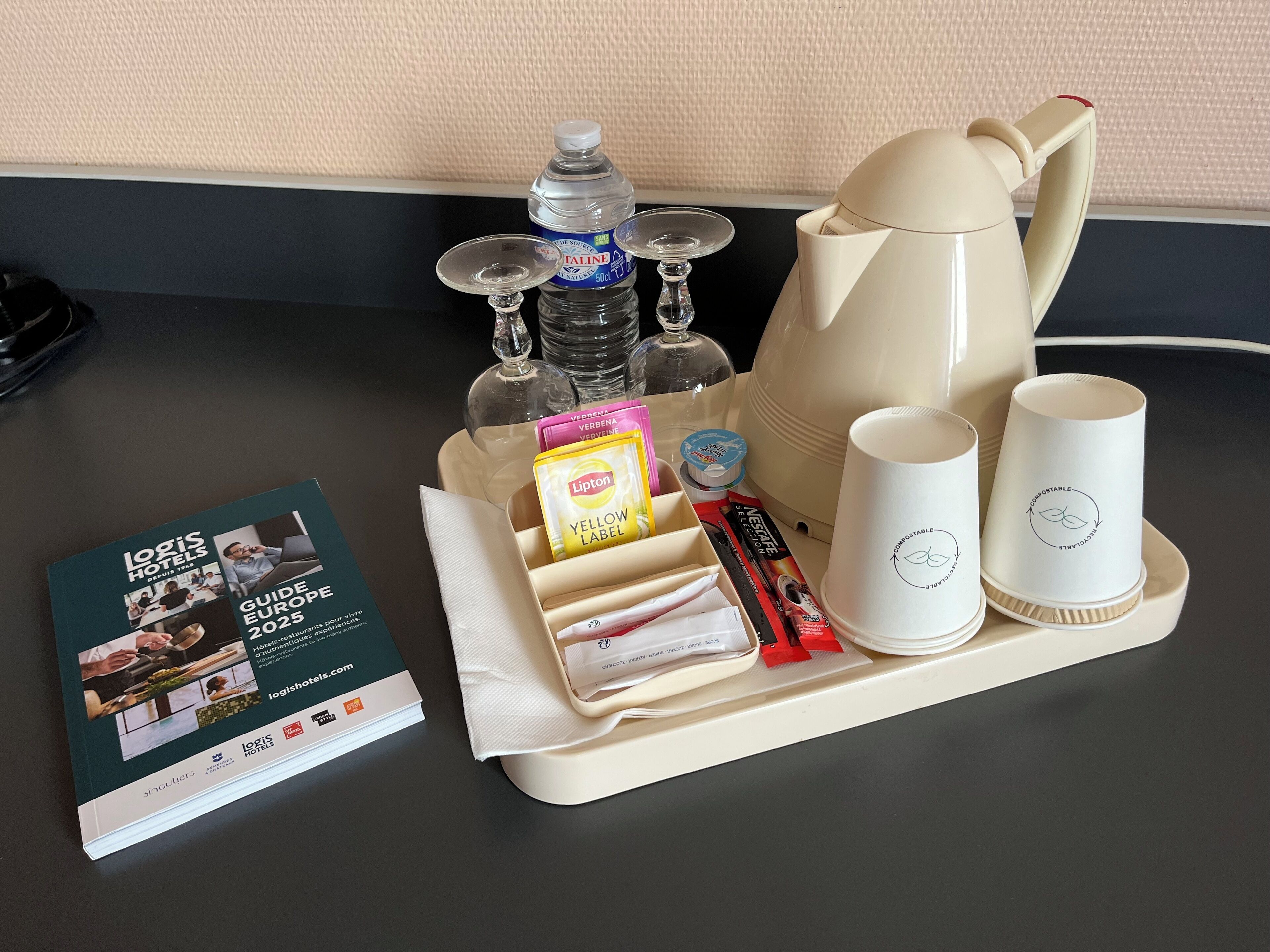 Un plateau contenant une bouteille d’eau, une bouilloire, deux tasses, une serviette en papier et un guide intitulé « Logis Hotels Guide Europe 2025 ».