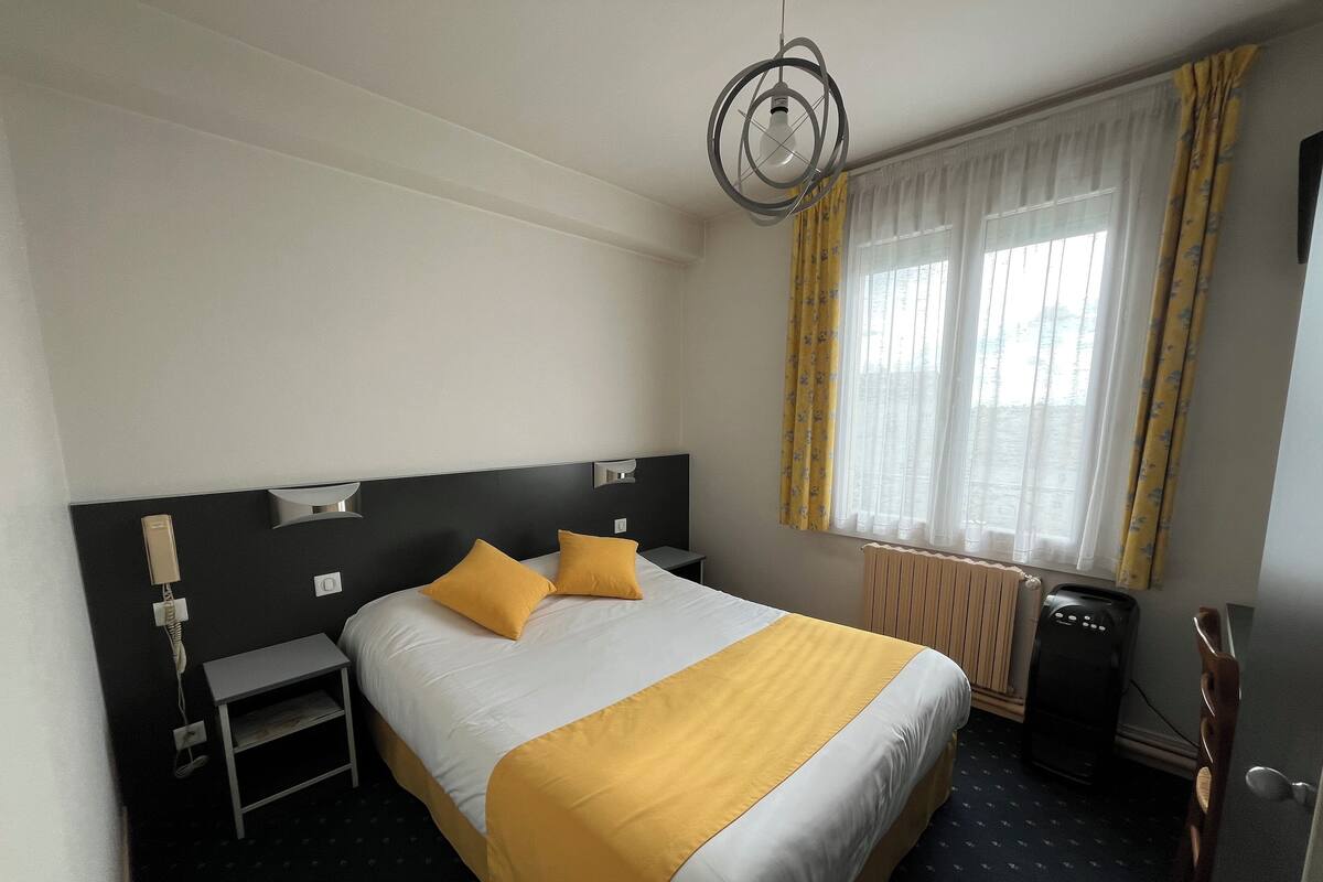 Une chambre d’hôtel comprenant un lit, des tables de chevet, un bureau, une chaise, un radiateur et une fenêtre avec des rideaux.