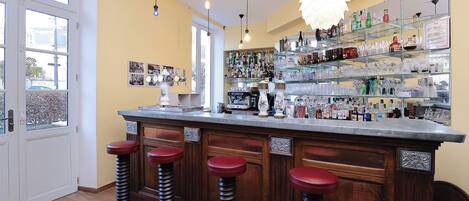 A bar area with a wooden counter, four stools, and a variety of bottles and glasses on shelves.