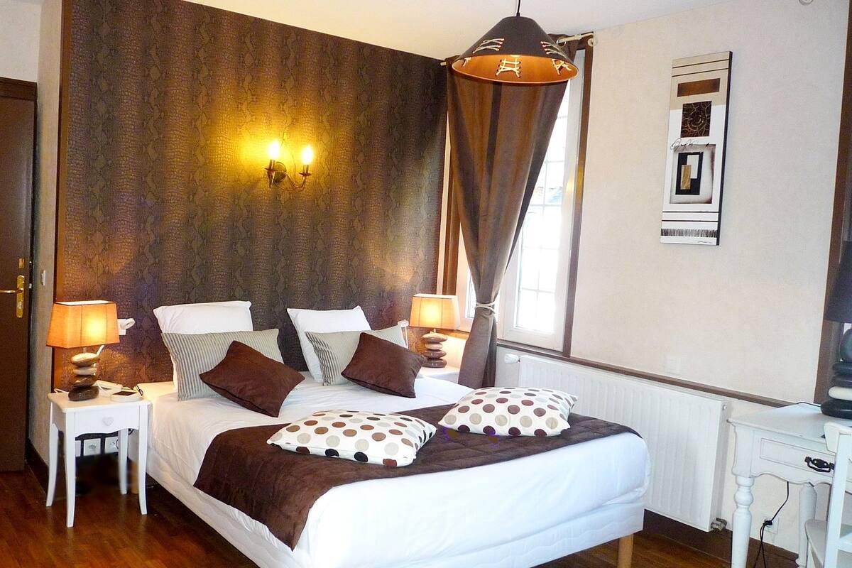 Habitación de hotel ordenada con una cama grande, dos mesitas de noche con lámparas, una lámpara de pared y un ventanal con cortinas.