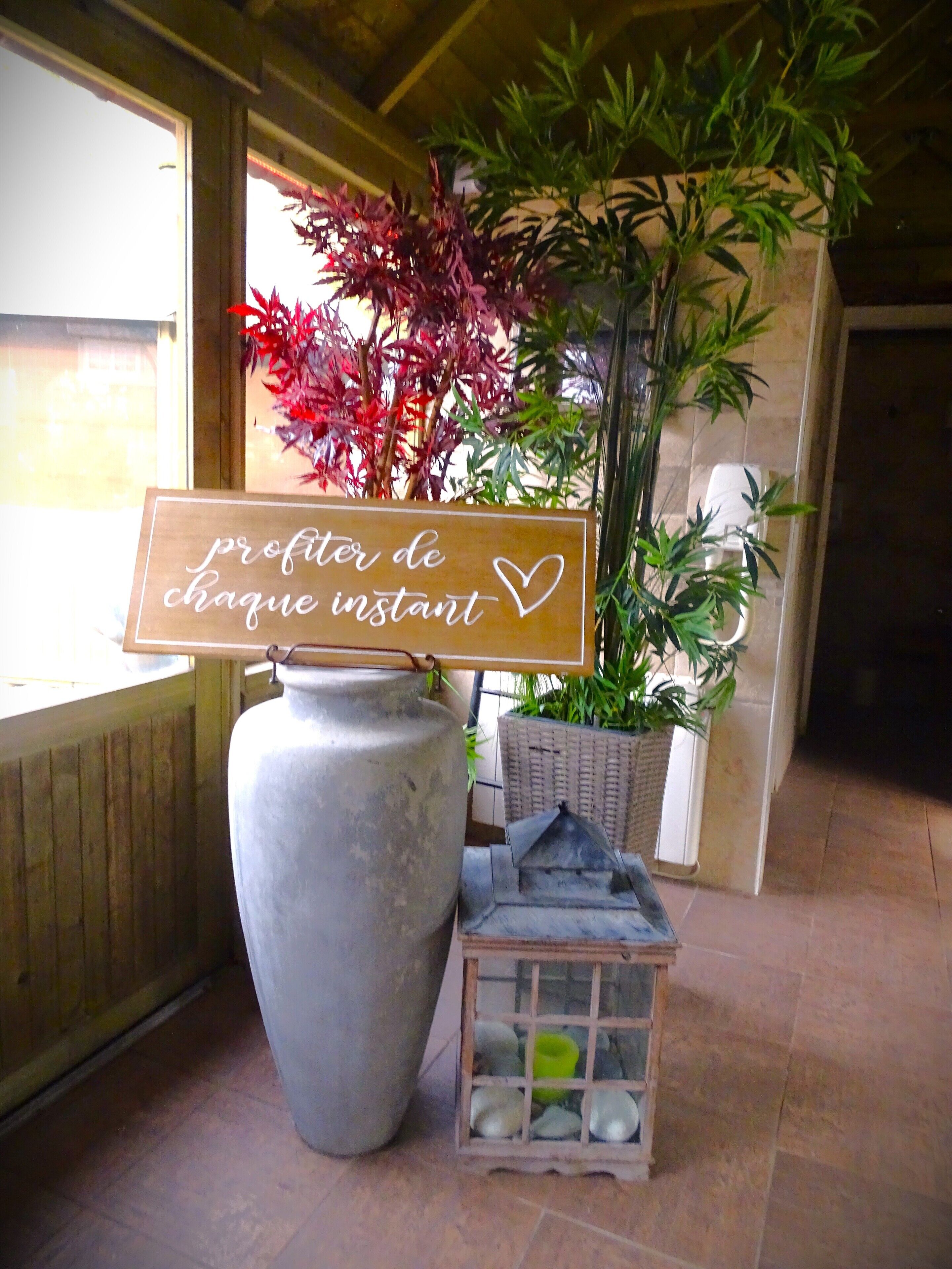 A wooden sign with text, a large vase, and potted plants in a room with tiled flooring.
