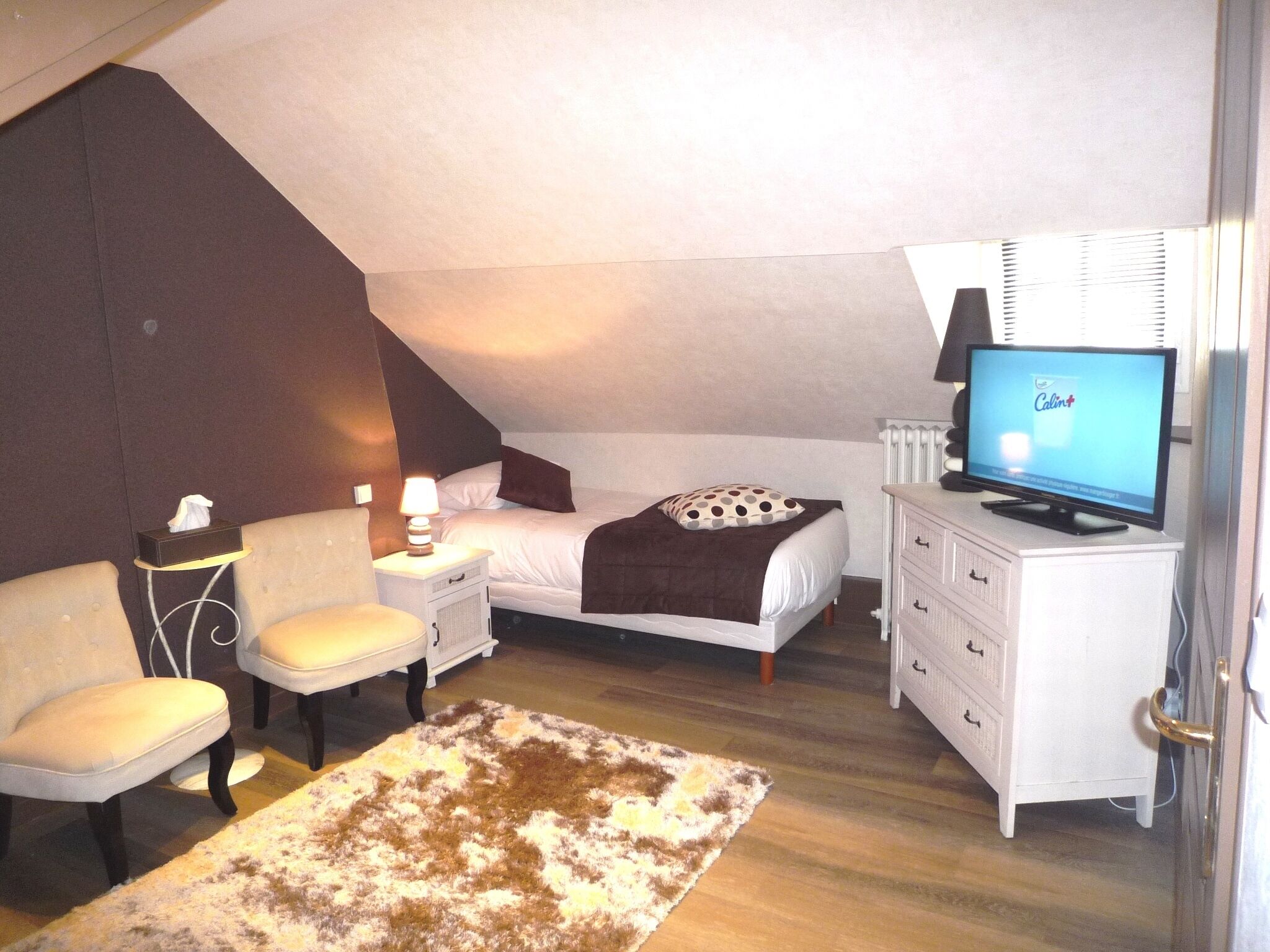 Ein Schlafzimmer mit einem Bett, einem Fernseher, einem Nachttisch, einem Sessel, einer Kommode und einem gemusterten Teppich.