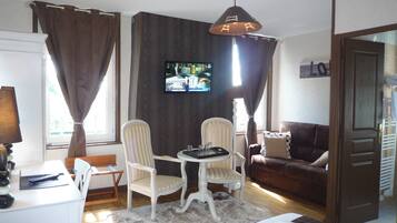 Una sala de estar con una mesa de comedor, dos sillas, un sofá, un televisor montado en la pared y una ventana con cortinas.