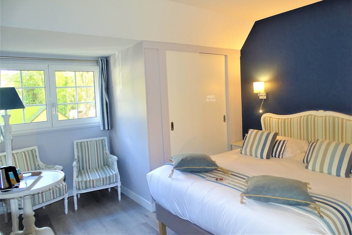 Una habitación de hotel con una cama, dos sillas, una mesita, una ventana con cortinas y una lámpara de pared.
