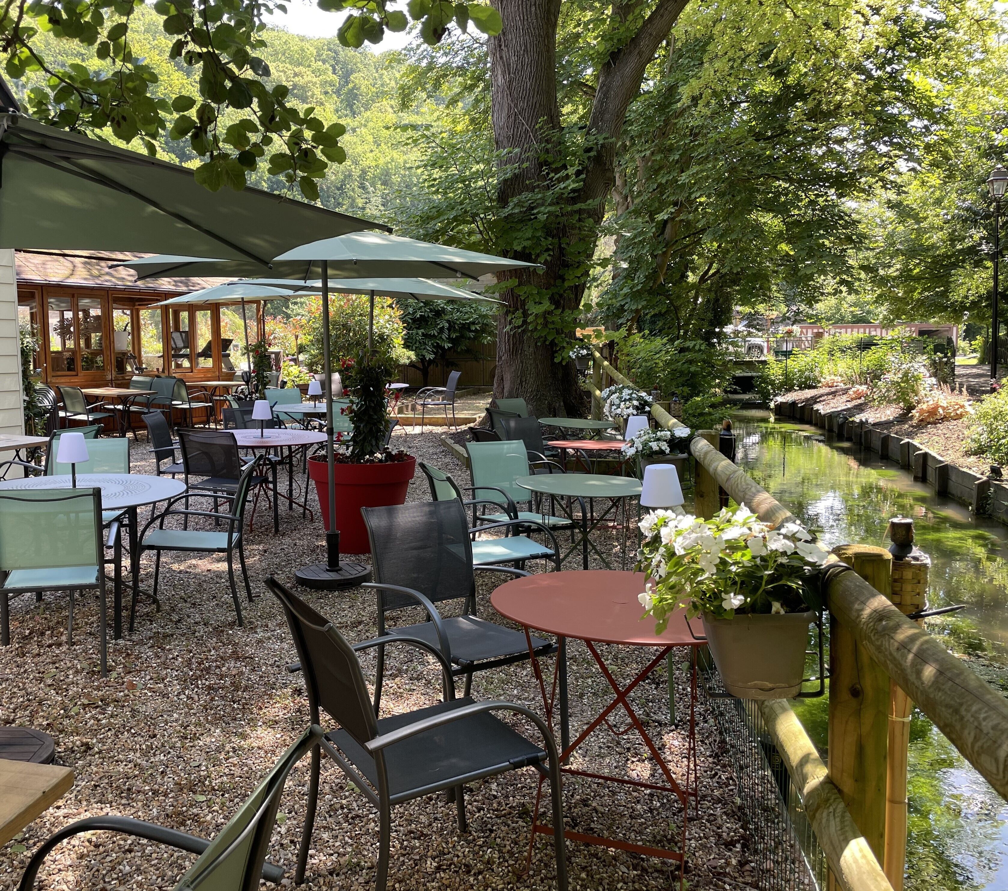 Een terras met tafels en stoelen, omringd door bomen en een waterpartij.