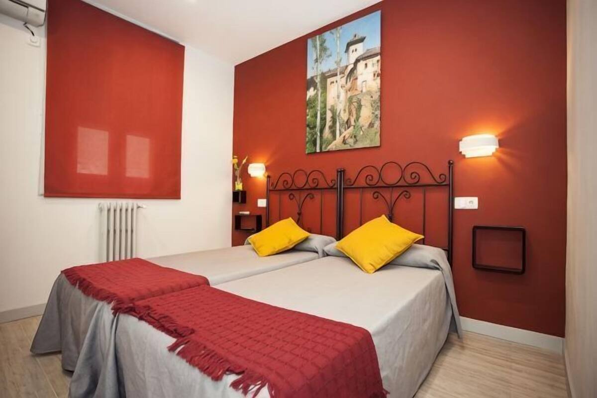 A double bed with red and white bedding, two yellow pillows, a red throw blanket, a wall-mounted light, and a framed picture above the bed.