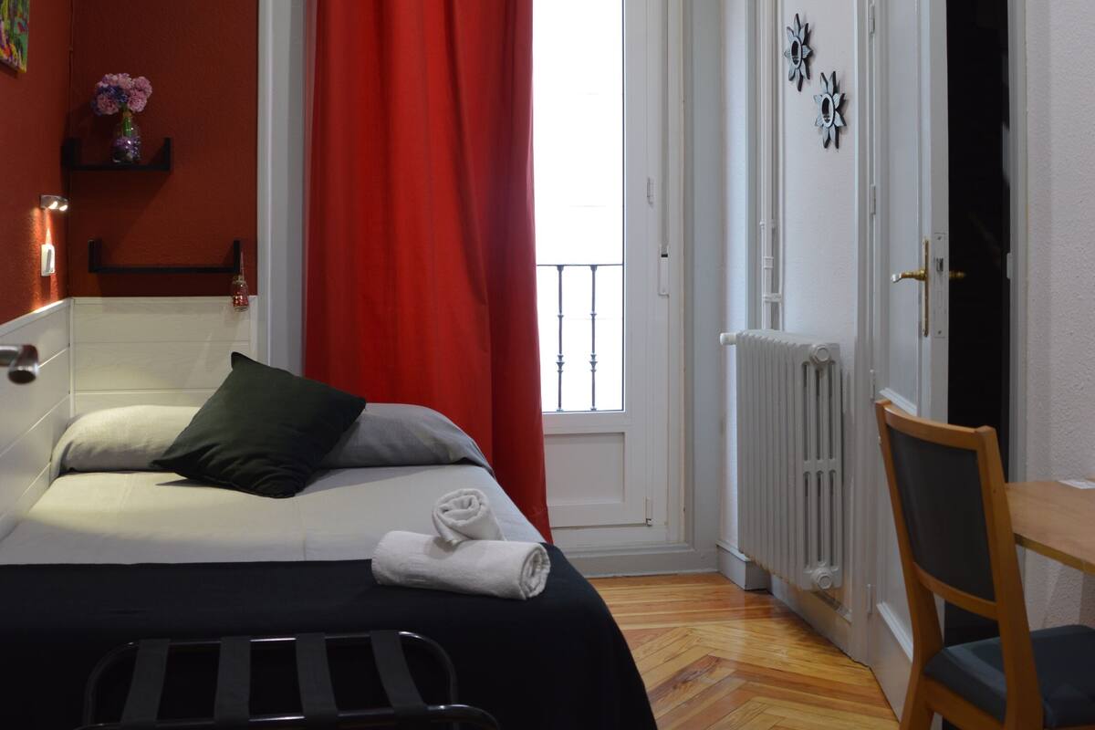 A bedroom with a bed, a chair, a radiator, and a window with red curtains.