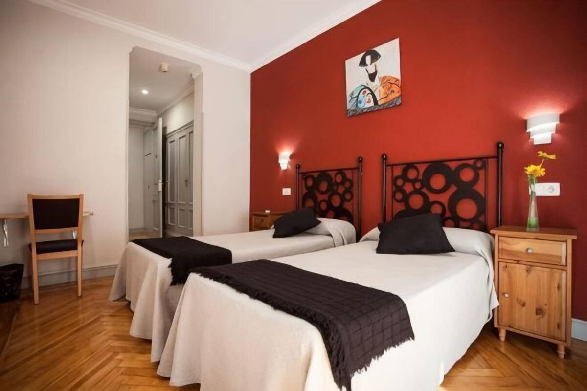 A room with two beds, a wooden chair, a nightstand, and a wall-mounted light fixture.
