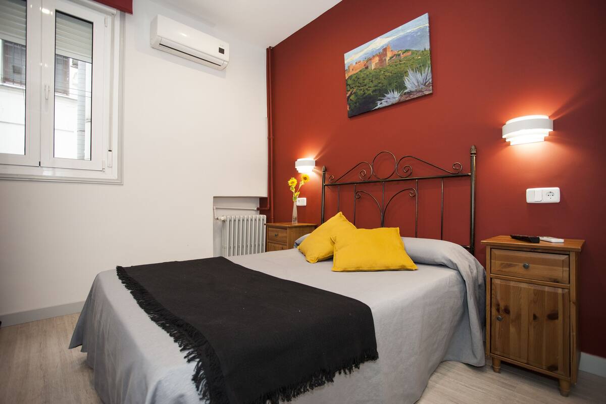A bedroom with a bed, a nightstand, a wall-mounted lamp, and a painting.