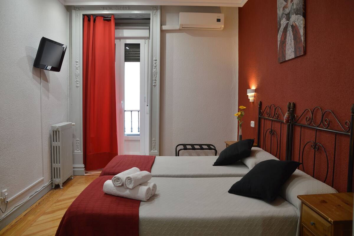 A hotel room with two beds, a TV mounted on the wall, a radiator, and a red curtain.