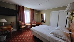 Une chambre d’hôtel avec un lit, deux chaises, un bureau, une lampe, un miroir et une fenêtre avec des rideaux.