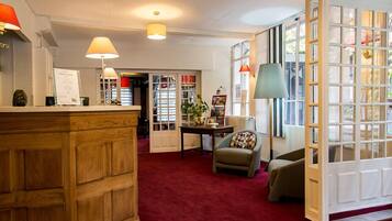 Eine Hotelhalle mit einer Rezeption, einem roten Teppich, einer Sitzecke mit Tisch und Stühlen sowie großen Fenstern mit Außenblick.
