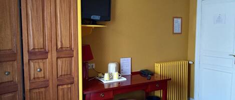 A hotel room with a wooden wardrobe, a desk with a chair, a bed, a television mounted on the wall, and a radiator.