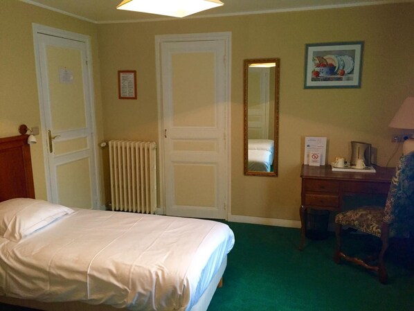 A hotel room with a bed, a desk with a chair, a radiator, a mirror, and a framed picture on the wall.