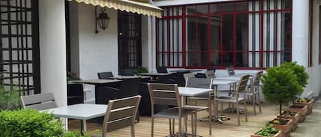 Un patio al aire libre con mesas y sillas de madera, rodeado de plantas en macetas.