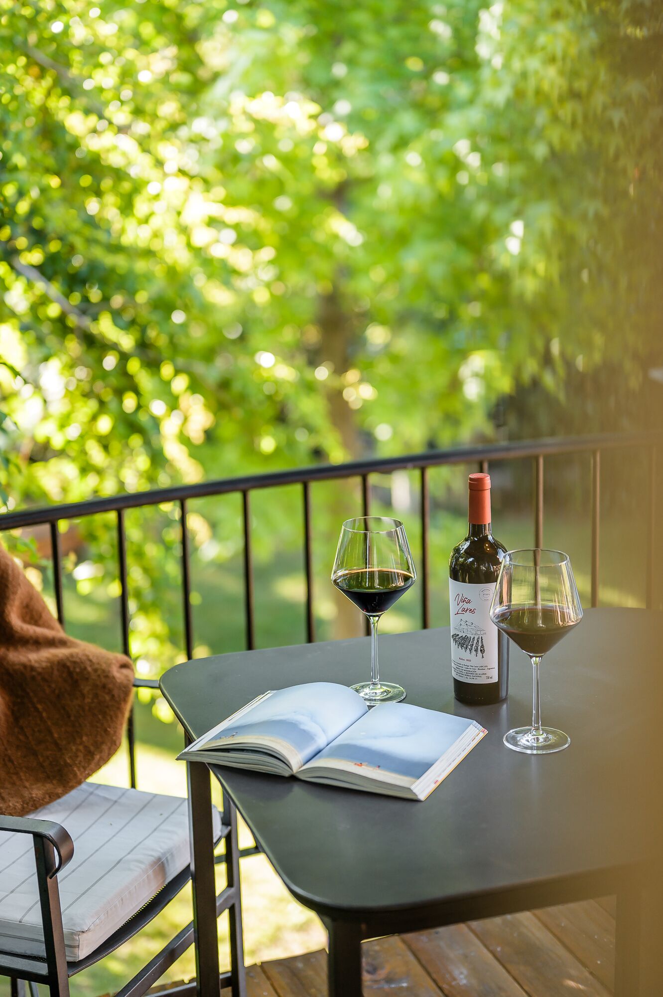 A table with a book, two wine glasses, and a wine bottle on a balcony with a railing.