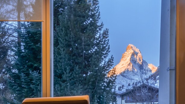 A modern room with a large window offering a view of a mountain and a hotel building. - Christiania Mountain & Spa (Zermatt)