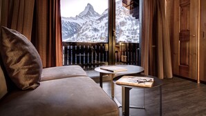 A modern room with a sofa, a small table with a book and glasses, and a large window with a view of snow-capped mountains. - Christiania Mountain & Spa (Zermatt)