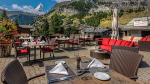 An outdoor dining area with tables and chairs, a red sofa, and a mountain view. - Christiania Mountain & Spa (Zermatt)