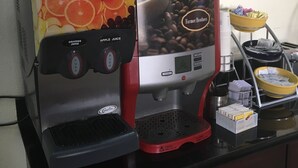 Une machine à café dotée de boutons pour préparer du jus d’orange et du jus de pomme, et une autre machine à café avec un compartiment permettant d’exposer les grains de café.