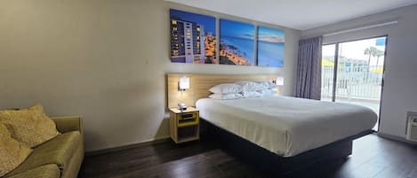 Ein Hotelzimmer mit einem großen Bett, einem Nachttisch, einer Lampe, einem Sessel und einem Fenster mit Blick auf den Strand und die Gebäude.