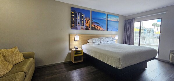 Un cuarto de hotel con una cama grande, una mesita de noche, una lámpara, una silla y una ventana con vista a la playa y edificios.