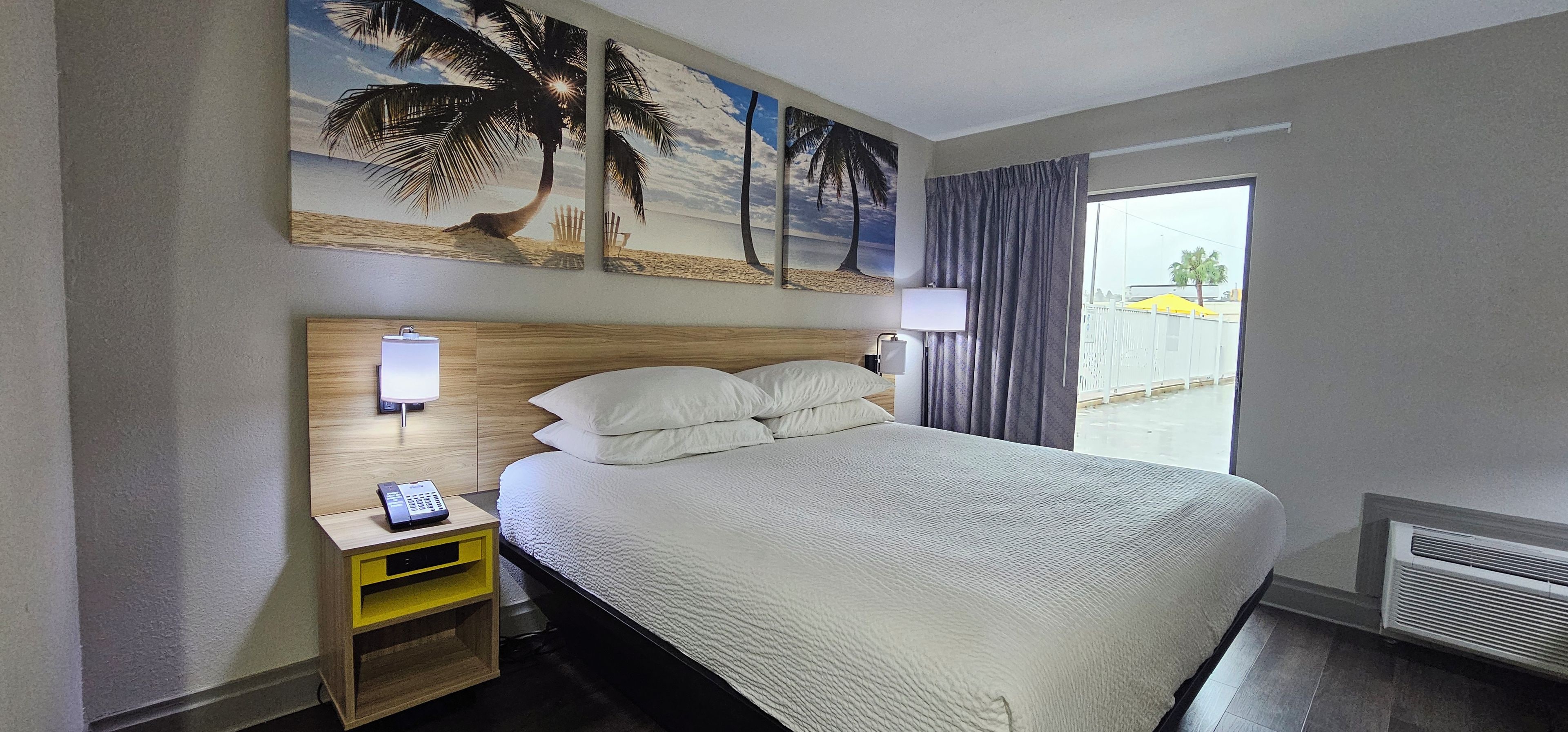 A hotel room with a bed, a nightstand with a phone, a wall-mounted lamp, and a painting of a beach scene.