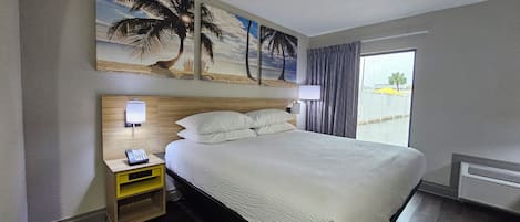 A hotel room with a bed, a nightstand with a phone, a wall-mounted lamp, and a painting of a beach scene.
