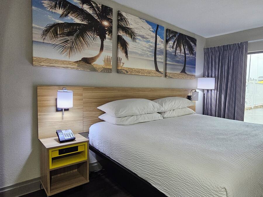 Una habitación de hotel con una cama, una mesita de noche con un teléfono, una lámpara de pared y un cuadro de una playa.