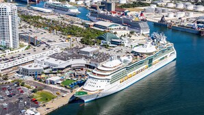 A large cruise ship docked at a port with a marina, terminal buildings, and a parking area.