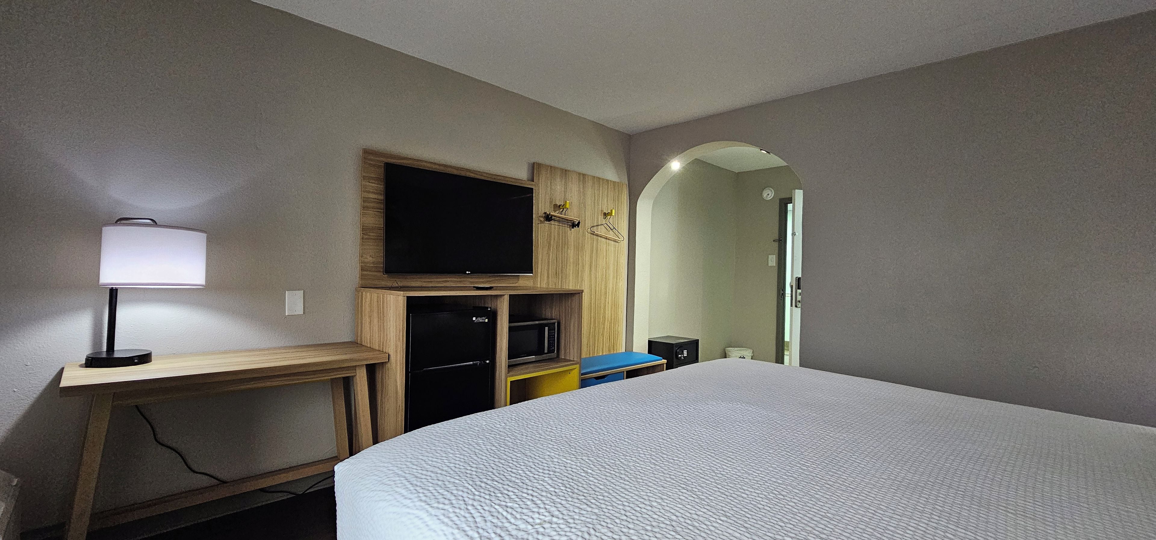 A bedroom with a bed, a TV mounted on a wooden cabinet, a desk with a lamp, and a microwave.
