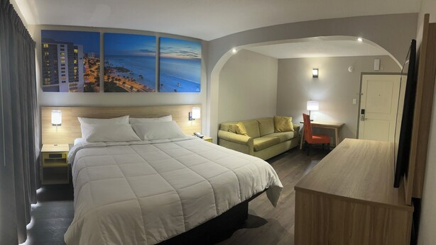 Une chambre d’hôtel avec un grand lit, un canapé, un bureau et une chaise. Les murs sont ornés de tableaux et la fenêtre offre une vue sur des immeubles et une plage.