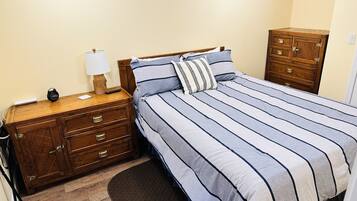 Un dormitorio con mesita de noche de madera, lámpara, cama con ropa de cama a rayas y cómoda de madera.